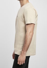 Boční pohled na muže stojícího s uvolněnými pažemi, oblečeného v béžovém tričku s krátkým rukávem a černých kalhotách na jednolitém pozadí.