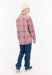 Persoon met de rug naar de camera, gekleed in een roze gestippeld jack, blauwe jeans, een beige muts en zwarte laarzen met beige zolen, op een witte achtergrond.