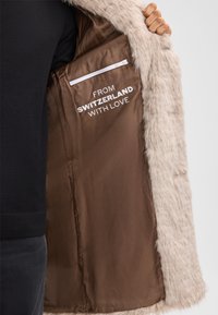Una persona tiene aperto un cappotto in faux fur beige, con foderato marrone, una tasca con zip bianca e la scritta "Dalla Svizzera con amore."