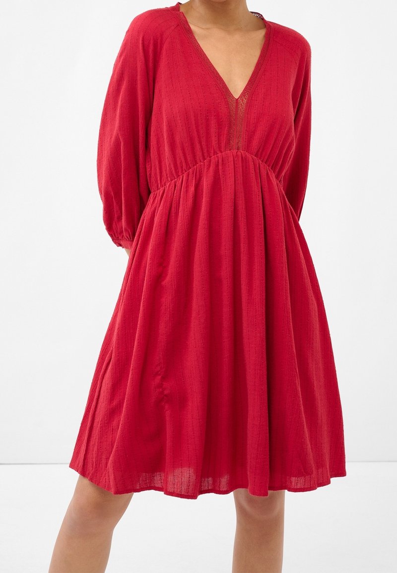 Femme portant une robe rouge mi-longue à manches longues et col en V, debout devant un fond blanc uni.