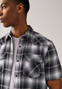 Kortärmad rutig skjorta i svart och grått med två bröstfickor, knappstängning framtill och en mjuk tygstruktur.