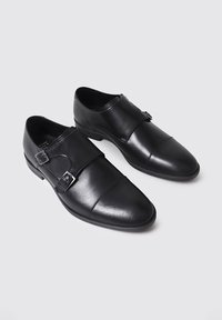 Zapatos de cuero negro con un acabado suave, que presentan dobles hebillas y una punta afilada. La suela es plana con una textura de relieve mínima.