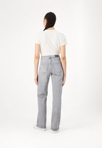 Ženska z zapletenimi lasmi stoji obrnjena proti ozadju v belem majici, svetlih sivo visokih kavbojkah in belih športnih čevljih na belem ozadju.