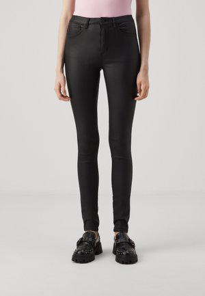 Vrouw die zwarte skinny jeans en zwarte loafers met studs draagt, staand tegen een effen lichte achtergrond.