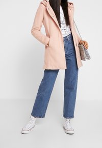 Cappotto rosa con cappuccio realizzato in tessuto morbido, con chiusura a zip e tasche laterali, abbinato a jeans blu a vita alta e sneakers bianche.