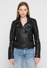 Zwarte leren motorjack met gequiltte schouderdetails, zilveren ritsen en een aangesloten ontwerp. Draagt over een lichtblauwe denimoutfit.