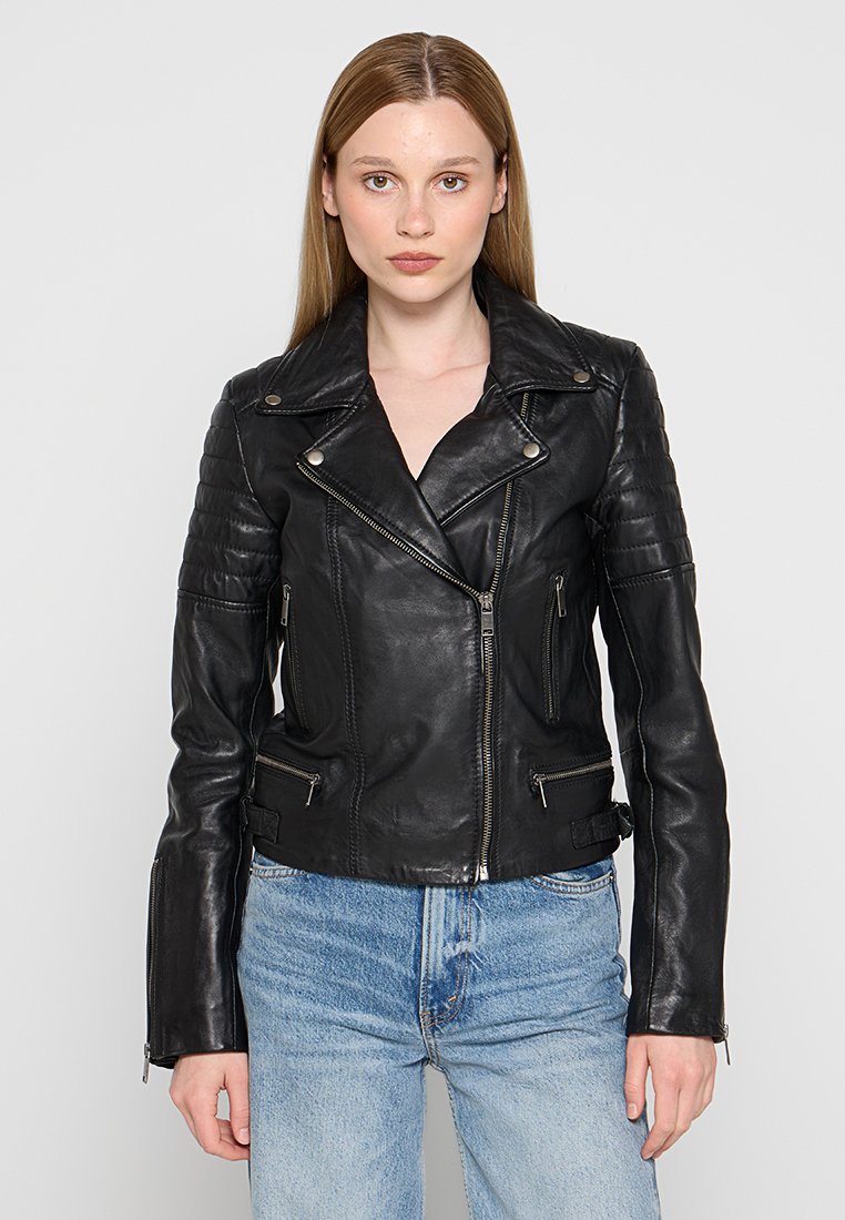 Zwarte leren motorjack met gequiltte schouderdetails, zilveren ritsen en een aangesloten ontwerp. Draagt over een lichtblauwe denimoutfit.