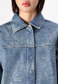 Denim jakna ima svetlo modro pranje z dekorativno vezenino iz rhinestones v vzorcu sončnega vzhoda in klasičen ovratnik.