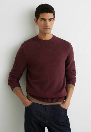 Mees, kes kannab bordo värvi kootud kampsunit beeži pikkade varrukatega särke ja tumelõuendilisi teksapükse, seisab käed taskutes valge seina taustal.