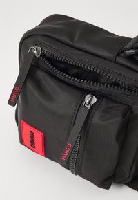 Borsa a tracolla nera in nylon con forma rettangolare, doppia cerniera e patch del logo rosso. Le cinghie includono tirazip brandizzati.