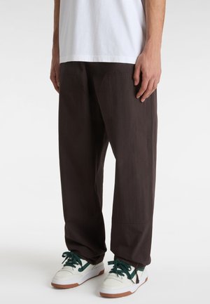 Bruine, lichte broek met een losse pasvorm, voorzien van voorzakken en een gestructureerde stof. Gecombineerd met witte sneakers en groene veters.