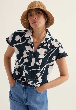 Frau mit einem beigefarbenen, gewebten Hut, einem schwarz-weiß geblümten Kurzarmhemd, goldener Halskette und Ohrringen sowie hoch taillierter blauer Jeans mit schwarzem Gürtel.