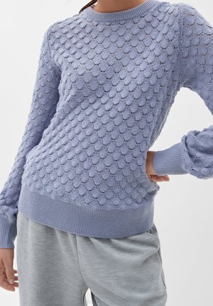 Femme portant un pull en maille texturée violet clair avec motif coquille et pantalon gris clair, main sur la hanche, sur fond uni.