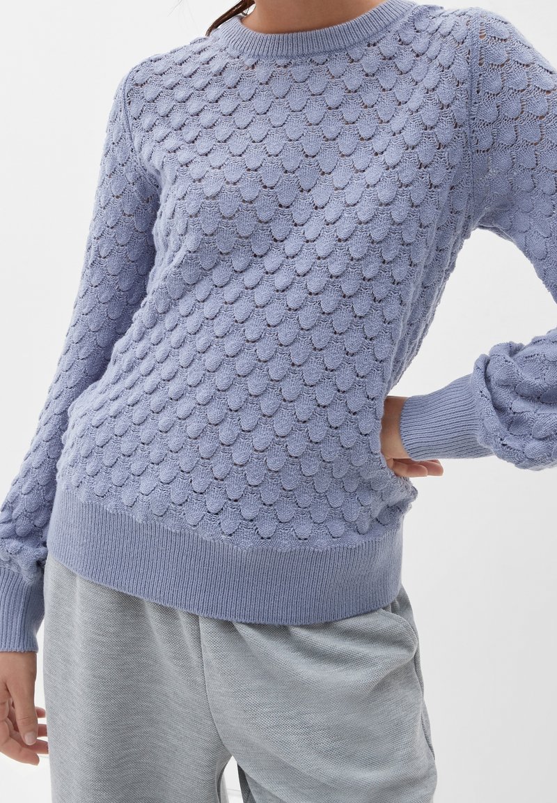 Femme portant un pull en maille texturée violet clair avec motif coquille et pantalon gris clair, main sur la hanche, sur fond uni.