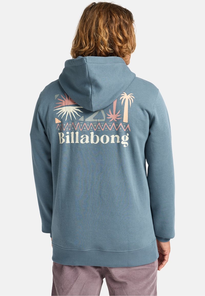 Personne aux cheveux mi-longs portant un sweat à capuche bleu avec un motif tropical pastel et le texte "Billabong" dans le dos.