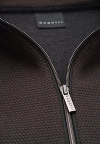 Reißverschlussjacke aus strukturiertem braunem Stoff mit geometrischem Muster, schwarer Innenauskleidung und silbernem Markenreißverschluss.