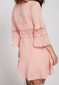 Vestido rosa texturizado con borde de encaje y mangas acampanadas. Presenta un dobladillo con volantes y una cintura ajustada. Tejido suave y ligero con un acabado arrugado.