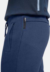 Navyblaue Sweatpants aus weichem Material mit einem Gummizugbund, Seitentasche und einem Markenpatch in der Nähe der Tasche.
