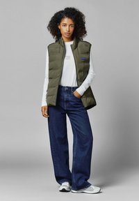 Olijfgroene puffer vest over een witte lange mouwen shirt, gecombineerd met losse donkerblauwe jeans en grijze sneakers. Eenvoudig ontwerp, geen patronen.