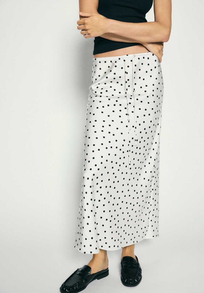 Femme portant une jupe midi blanche à pois noirs, un top noir sans manches et des mocassins noirs sans lacets, debout devant un fond uni.