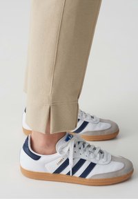 Personne portant un pantalon beige avec des fentes latérales et des baskets blanches Adidas Samba avec des bandes bleu marine et des semelles en gomme.