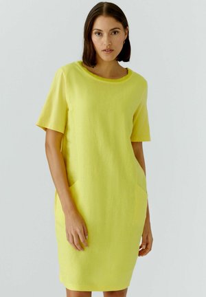 Femme aux cheveux foncés mi-longs portant une robe jaune vif à manches courtes et longueur genoux avec des poches devant, debout devant un fond uni.