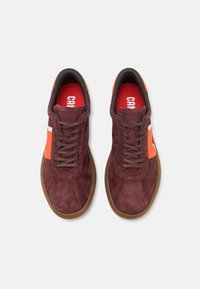 Mörkröda mockaskor med orange accenter, rund tå, platt brun sula och bruna skosnören. Logotyp synlig på tungan och sidan.