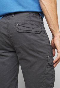 Primo piano di una persona che indossa pantaloni cargo grigio scuro con tasche a patta e una camicia blu, con una mano che riposa vicino alla tasca.