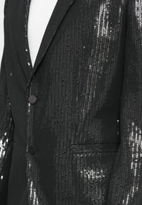 Zwarte blazer met een sequined textuur, twee voorknopen, een reverskraag en een zijzak. De stof heeft een gladde afwerking met reflecterende accenten.