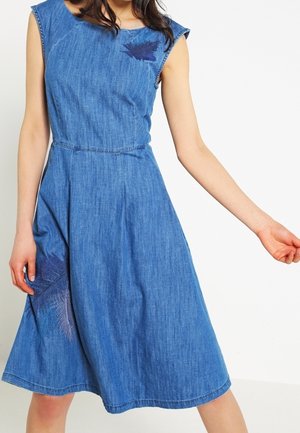 Sleeveless blue denim midi dress with subtle dark feather prints on chest and skirt, worn by a person with one arm extended.