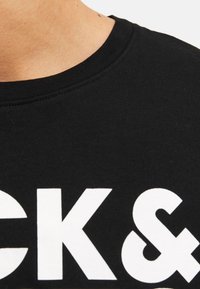 Black cotton T-shirt with a crew neck, featuring large white text that reads "CK&" in bold typography. Close-up view of neckline and print.