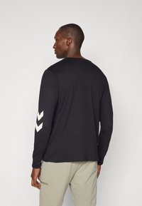 Juoda ilgomis rankovėmis marškiniai su apvaliu kaklu ir baltais chevrono raštais ant rankovių. Audinys atrodo lygus, stilius sukurtas atsipalaidavusiam pritaikymui.