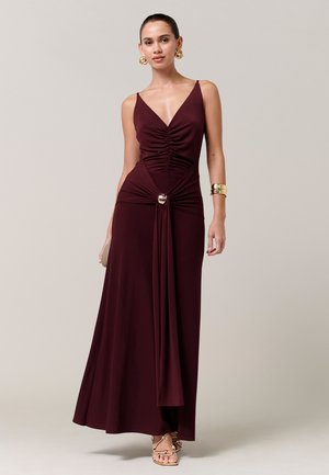 Mujer con un vestido maxi sin mangas de color burdeos intenso con fruncido en la parte delantera, detalle de anillo metálico, pendientes dorados, pulsera y sandalias de tiras.