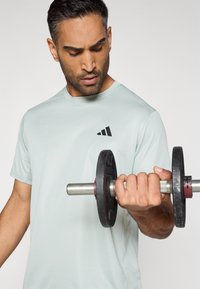 Siva športna majica s kratkimi rokavi, z črnim logotipom. Oseba drži črno utež z kovinskim ročajem. Prikaz iz bližine.