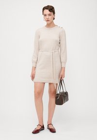 Strikket beige kjole med lange ermer, innsnevret midje med bånd og knappdetaljer på skulderen. Kombinert med brun håndveske og bordeaux sko.
