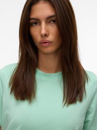 Jauna moteris su tiesiais rudos spalvos plaukais, dėvi šviesiai žalią marškinėlį, neutralaus veido išraiška, stovinti priešais paprastą baltą foną.