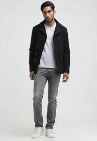Man in een zwarte pea coat, witte V-hals t-shirt, grijze jeans en witte sneakers, staande tegen een effen lichte achtergrond.