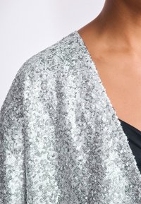 Silberner Paillettenmantel mit glatter, schimmernder Oberfläche; verfügt über einen lockeren, fließenden Fall und einen V-Ausschnitt, getragen über einem schwarzen Oberteil.