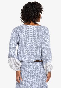 Blauwe blouse met een patroon, lange, losse mouwen en een brede halslijn. Bevat een bloemenmotief en een contrasterende witte mouwzoom.