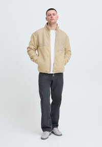 Giacca in pile beige con una parte superiore liscia, colletto alto e chiusura con zip. Abbinata a pantaloni in denim scuri e scarpe grigie chiare.