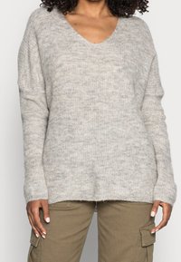 Maglione oversize grigio chiaro con scollatura a V, maniche lunghe e motivo a maglia texturizzata. Indossato con pantaloni cargo color khaki.