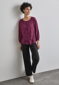 Luźna bluzka w różowo-fioletowy wzór z długim rękawem. Zestawiona z czarnymi dżinsami o wysokim stanie i białymi trampkami.