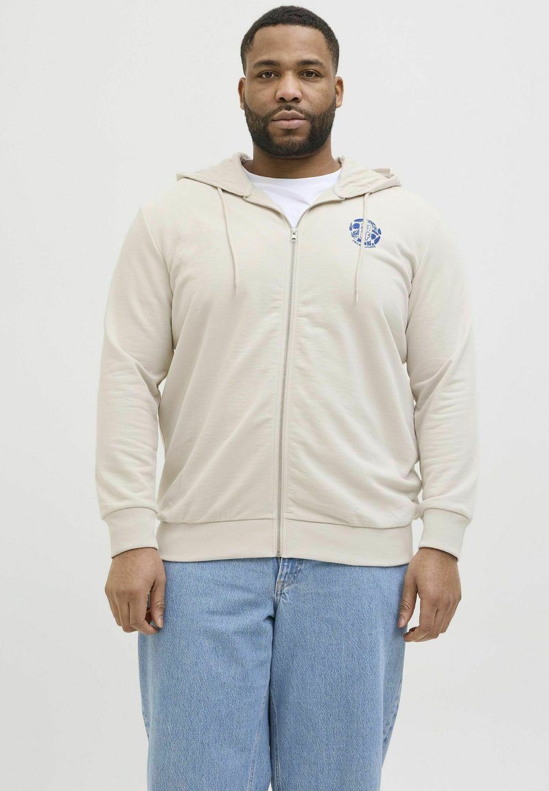 Beige zip-up hoodie med snøre og et brystlogo, lavet af blødt stof. Båret over en hvid skjorte og parret med lyseblå jeans.