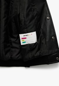 Interior de chaqueta negra acolchada con textura suave. Presenta una etiqueta de cuidado con secciones coloridas para nombre, clase y detalles de contacto.
