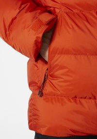 Červená péřovka s boční kapsou na zip a nastavitelnými šňůrkami. Hladká, lehká tkanina s prošívanými částmi.