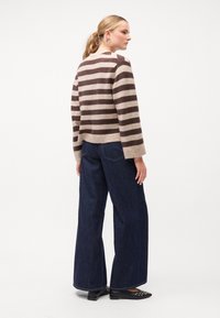 Καφέ και μπεζ ριγέ πλεκτό πουλόβερ με στρογγυλή λαιμόκοψη, συνδυασμένο με τζιν παντελόνια ψηλής μέσης σε σκούρο μπλε χρώμα και μαύρα επίπεδα παπούτσια.