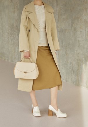 Person trägt beige Trenchcoat, beige gerafftes Oberteil, senfgelben Rock, weiße Schuhe mit Holzabsatz und hält eine cremefarbene Handtasche.