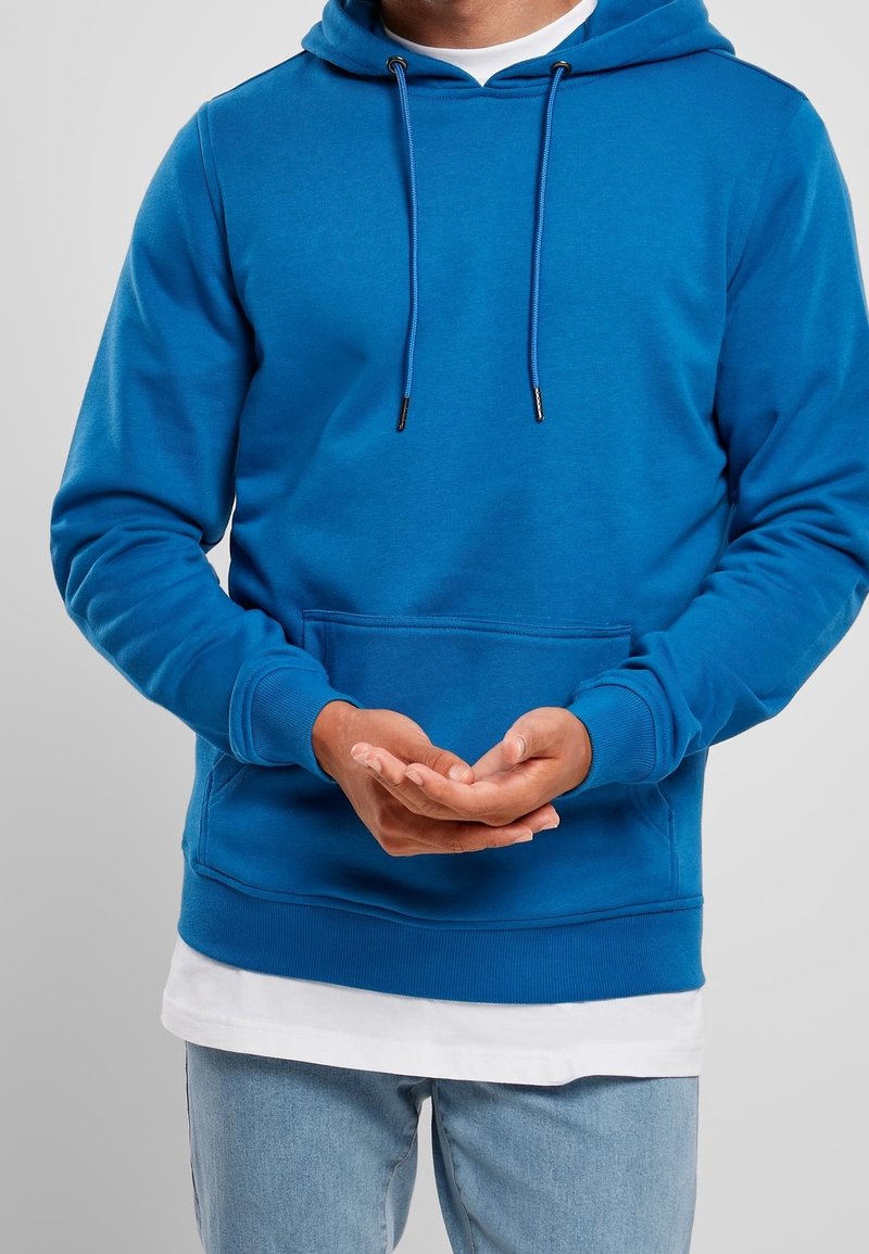 Homme portant un sweat à capuche bleu et un jean bleu clair, les mains légèrement jointes devant un fond gris.