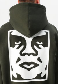 Donkergroene hoodie met een groot wit grafisch ontwerp van een gestileerd gezicht op de achterkant. De stof lijkt zacht met een gladde textuur.