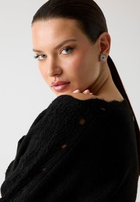 Maglione nero in tessuto testurizzato con fori circolari, dotato di scollatura ampia e materiale in maglia morbida. Modella che indossa orecchini a bottone argento, rivolta verso la fotocamera.